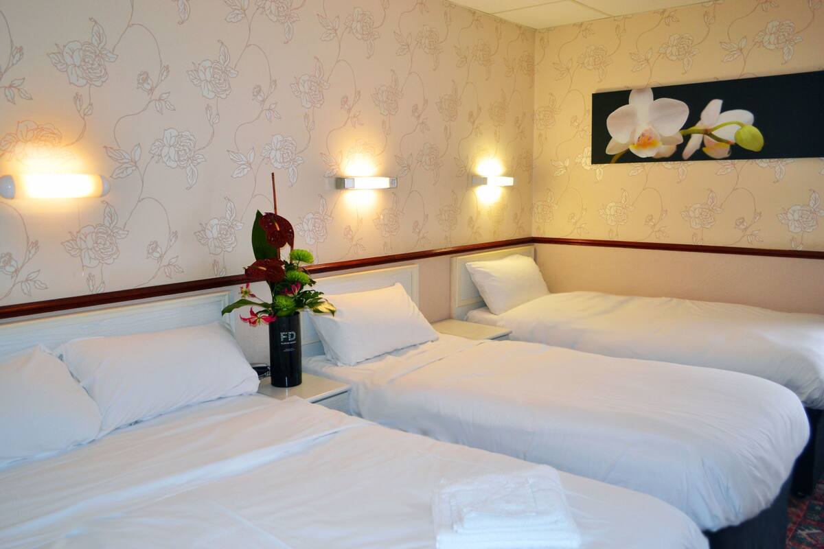 A hotel room with two single beds, a nightstand with a vase of flowers, and wall-mounted lights.