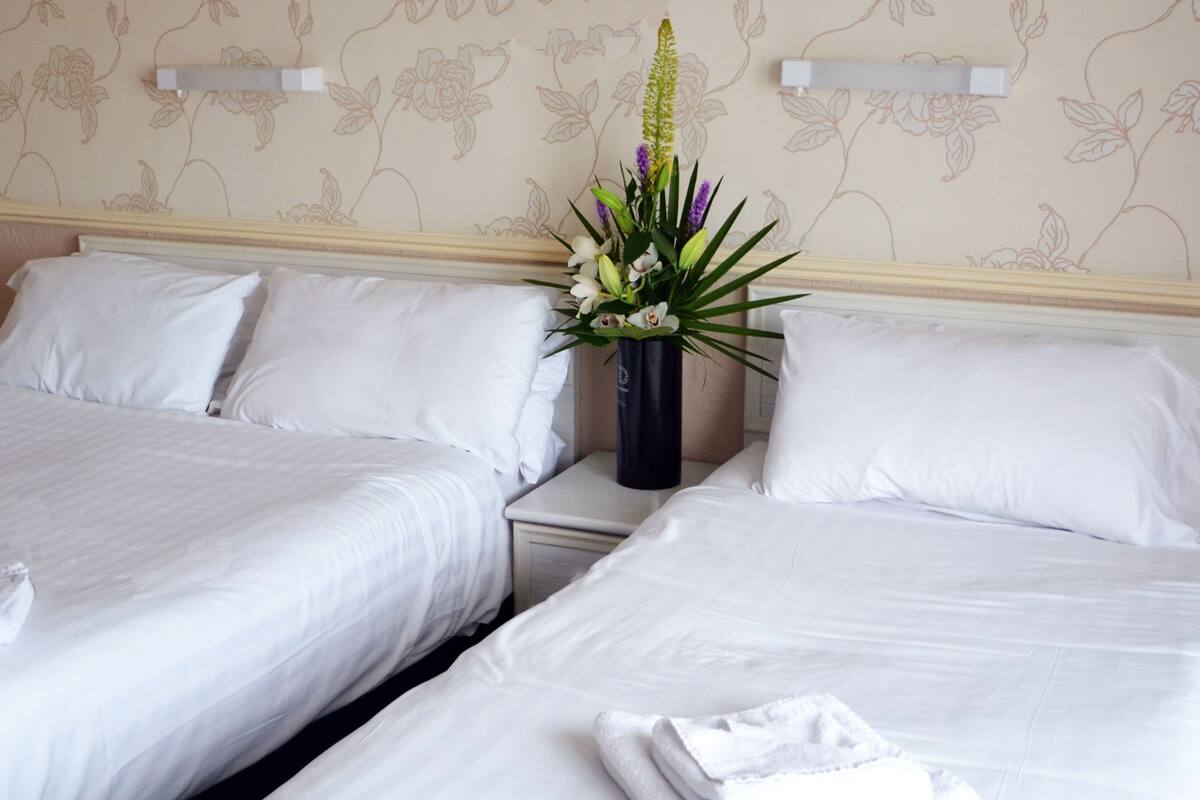 A hotel room with two beds, white linens, a vase with flowers, and a floral wallpaper design.