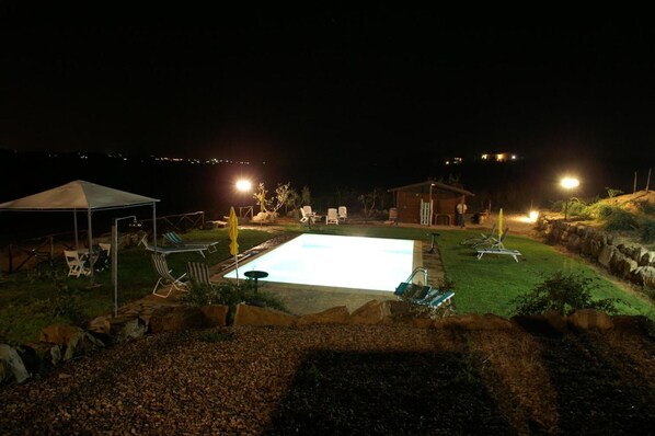 A nighttime outdoor swimming pool area with lounge chairs and a covered seating area.