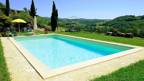 Ein rechteckiger Pool mit Steinumrandung, umgeben von einer Rasenfläche mit einem gelben Sonnenschirm und Liegestühlen.
