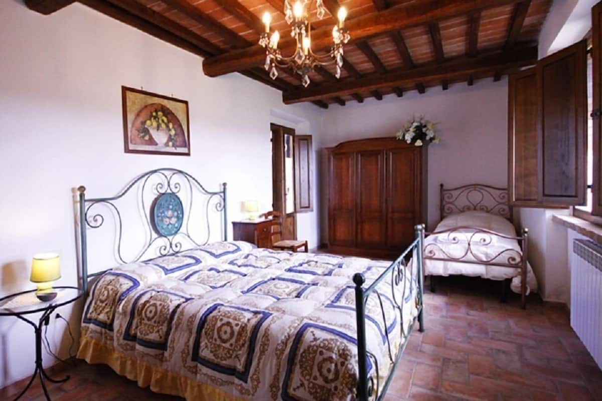 A bedroom with a large bed, a smaller bed, a wooden wardrobe, a table with a lamp, and a chandelier.