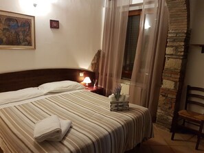 A hotel room with a bed, a nightstand with a lamp, a framed picture on the wall, a window with curtains, and a stone wall. - Agriturismo Il Giogo (Rosignano Marittimo)