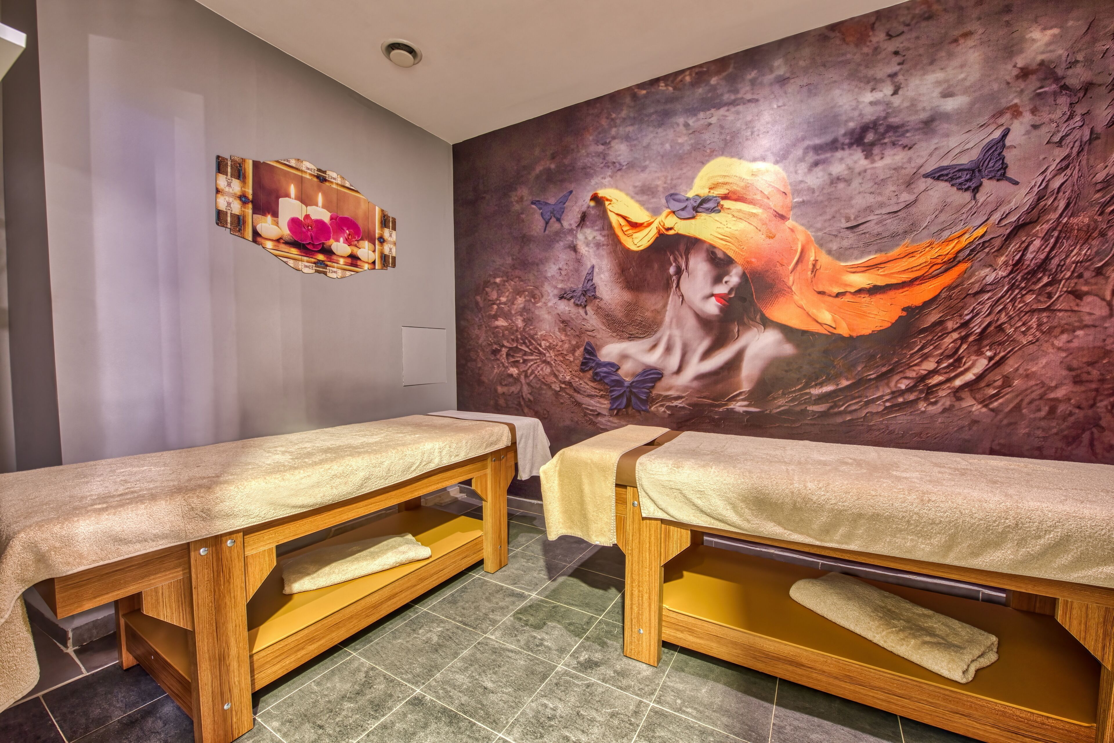 A spa room with two wooden massage tables, a large mural on the wall, and a small wooden shelf with candles and flowers.