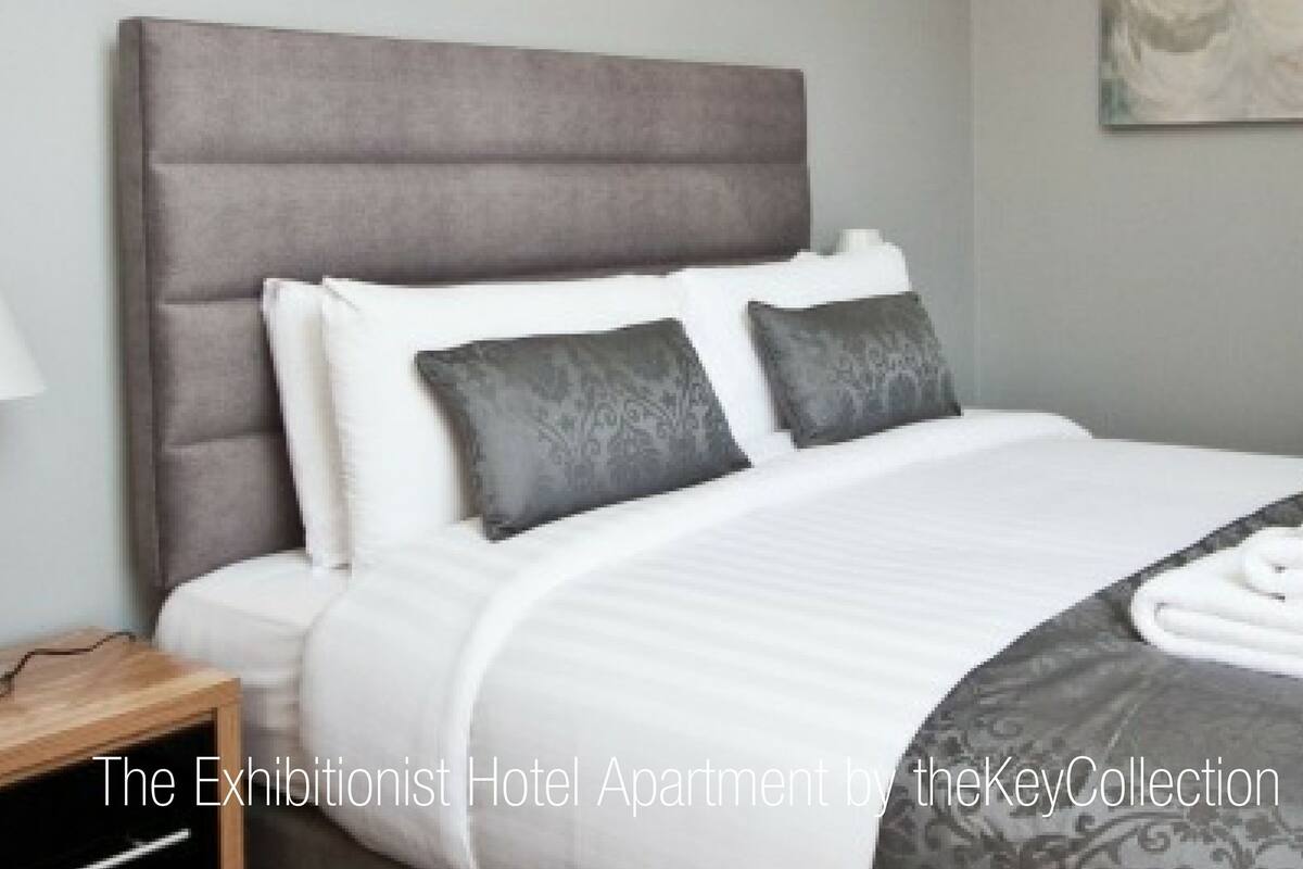 A hotel bedroom with a large bed, grey headboard, white bedding, patterned throw, and neatly folded towels.