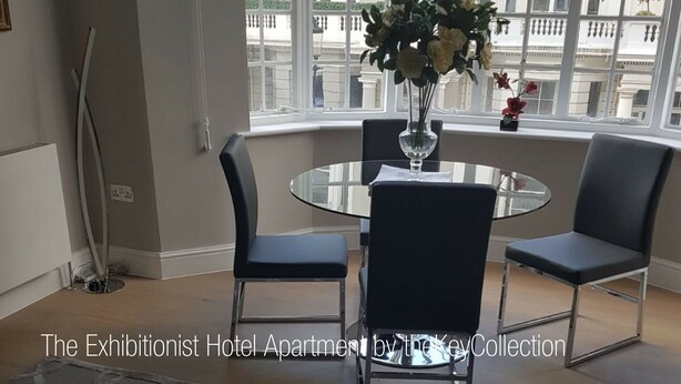 Een hotelappartement met een ronde glazen tafel, twee zwarte stoelen, een bloemenvaas en uitzicht op gebouwen door grote ramen.