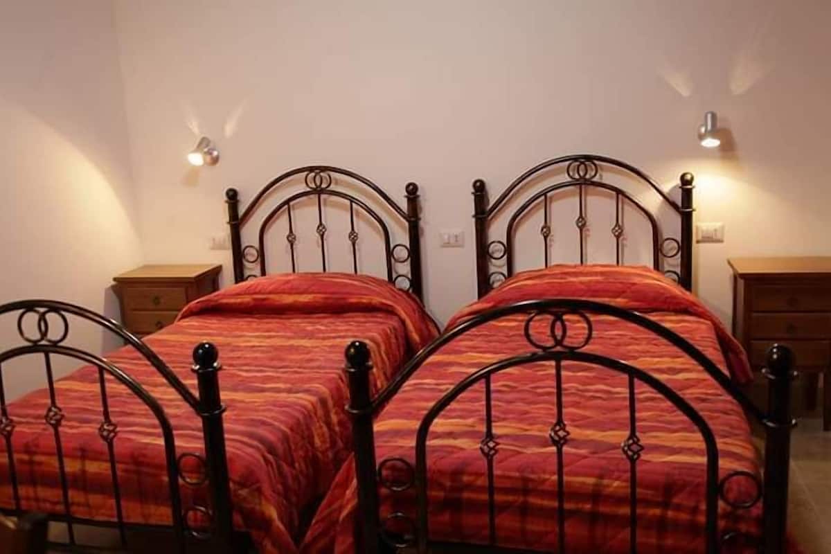 A room with two metal-framed beds with red bedding, a wooden nightstand, and a wall-mounted light fixture.