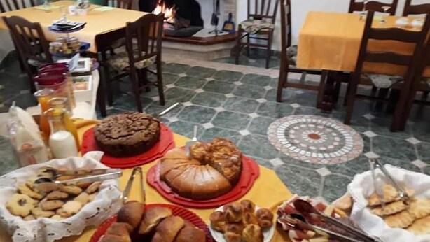A dining table set with various baked goods and snacks, surrounded by chairs and a fireplace. - B&B Gennargentu (Fonni)