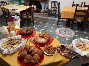 A dining table set with various baked goods and snacks, surrounded by chairs and a fireplace. - B&B Gennargentu (Fonni)