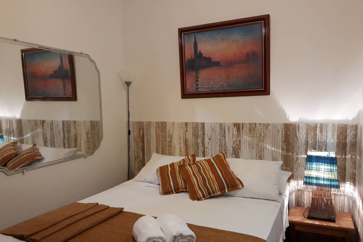 A hotel room with a bed, two paintings, a bedside lamp, and a small wooden table.