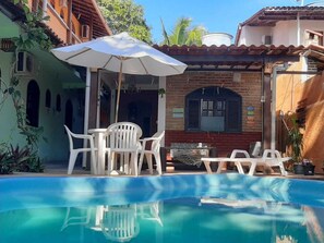 A swimming pool with white plastic chairs and an umbrella, surrounded by a brick building with a tiled roof. - Pousada Rota Tropicana (Porto Seguro)