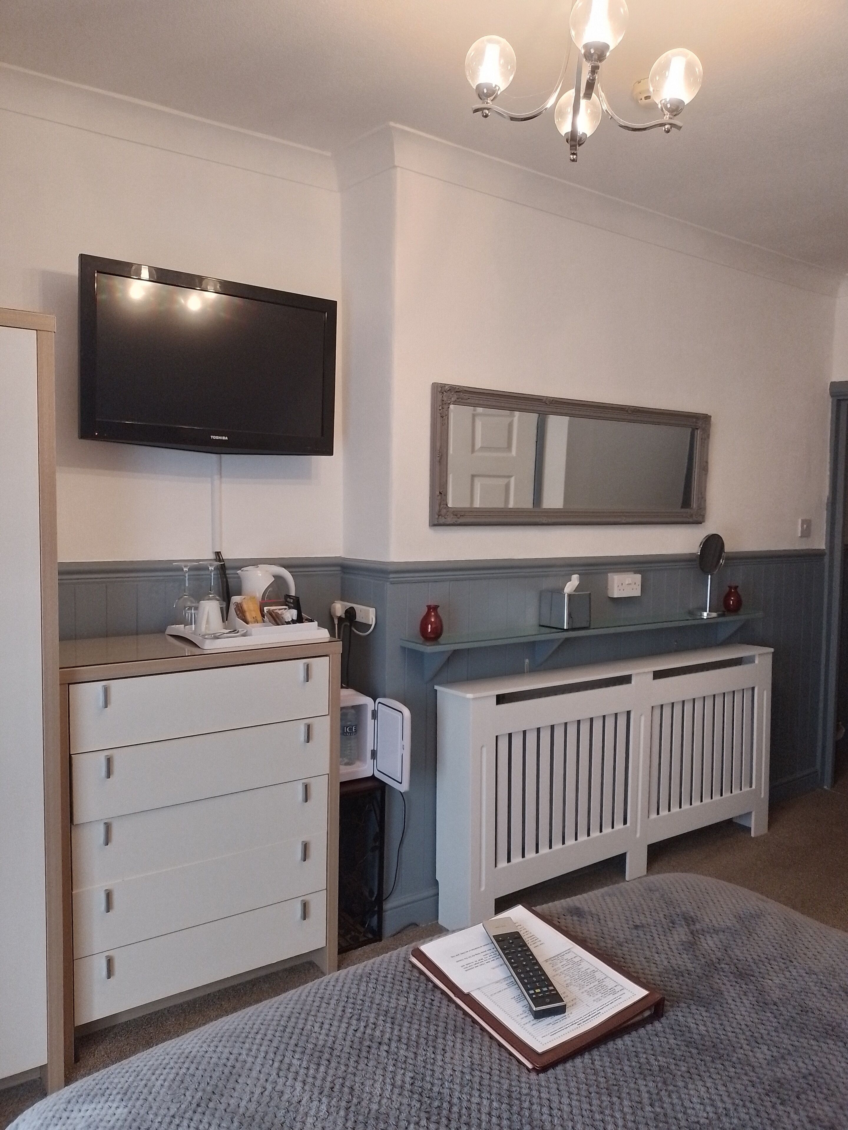 Een kamer met een wandgemonteerde flatscreen-tv, een witte dressoir met lades, een glazen plank met decoratieve objecten, een witte radiator, en een gebreide vloerkleed.