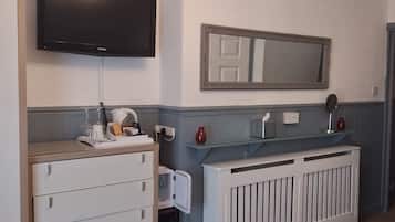 Una habitación con un televisor de pantalla plana montado en la pared, un aparador blanco con cajones, un estante de vidrio con objetos decorativos, un radiador blanco y una alfombra de punto en el suelo.