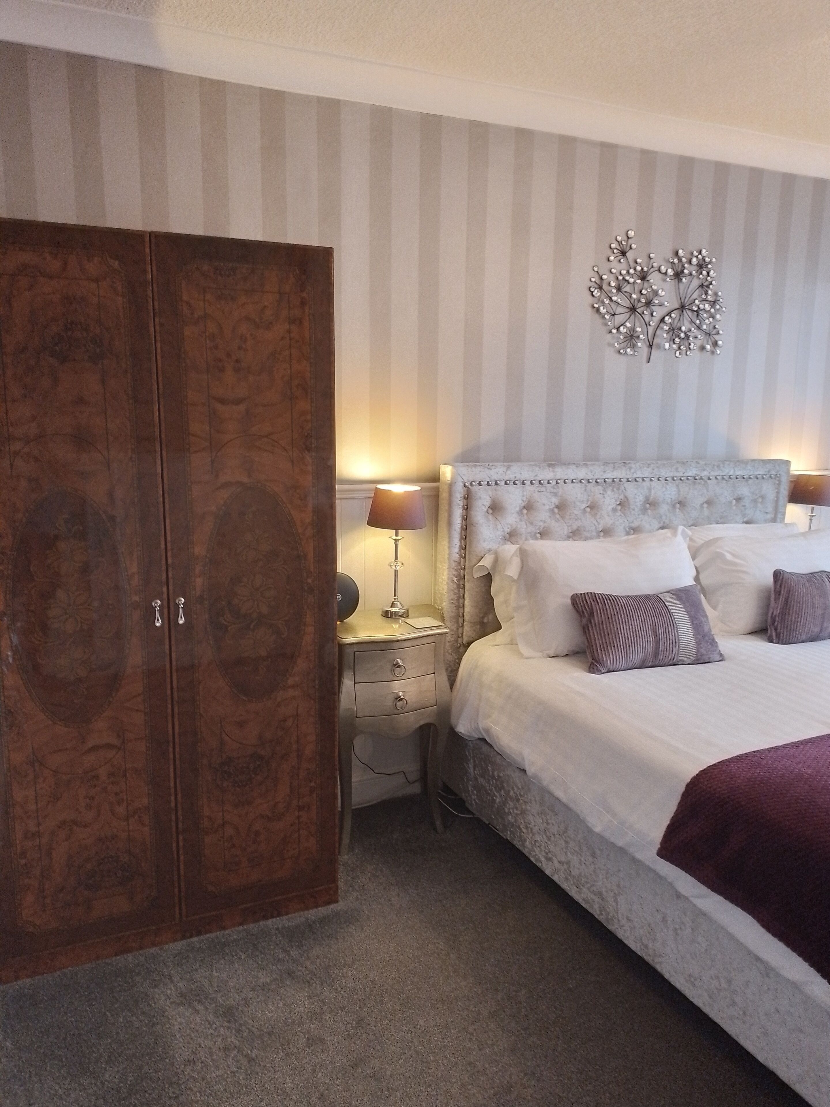 A bedroom with a large wooden wardrobe, a bed with white bedding and decorative pillows, a bedside lamp, and a wall-mounted decorative piece.
