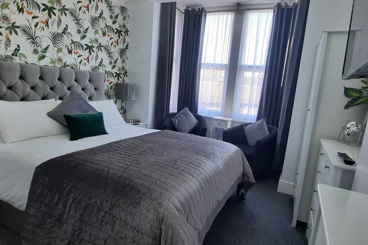 A hotel room with a tufted headboard bed, a grey quilted bedspread, and a floral wallpaper. There is a bedside table with a lamp, a small desk with a chair, and a large mirror with a decorative frame.