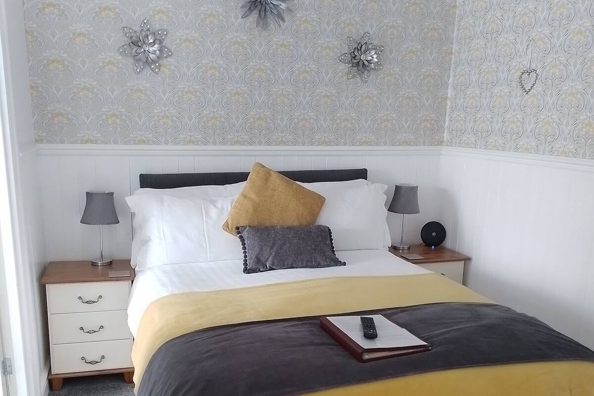 A neatly made bed with a yellow and white bedspread, two pillows, and a remote control on the bed. There are two bedside tables with lamps, a wall clock, and a decorative wall piece above the bed.