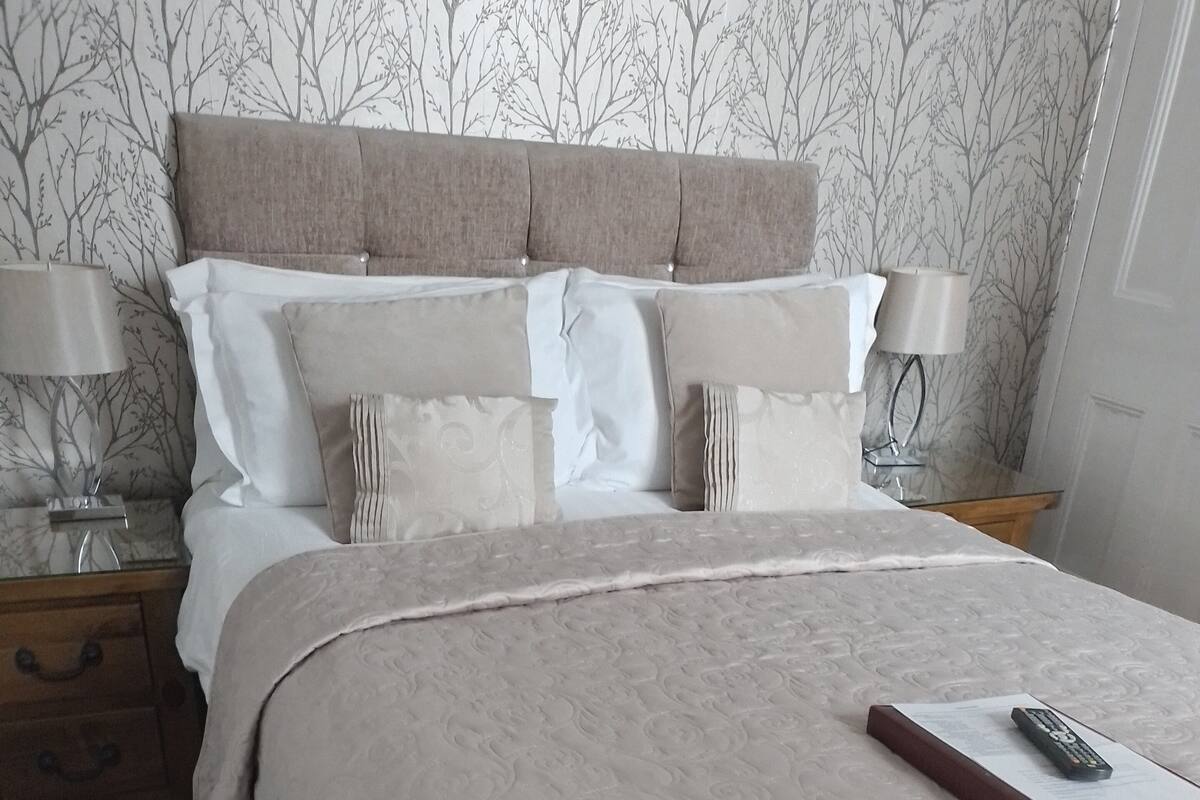 A neatly made bed with a beige comforter and pillows, a wooden nightstand with a lamp, and a wall with a tree pattern.