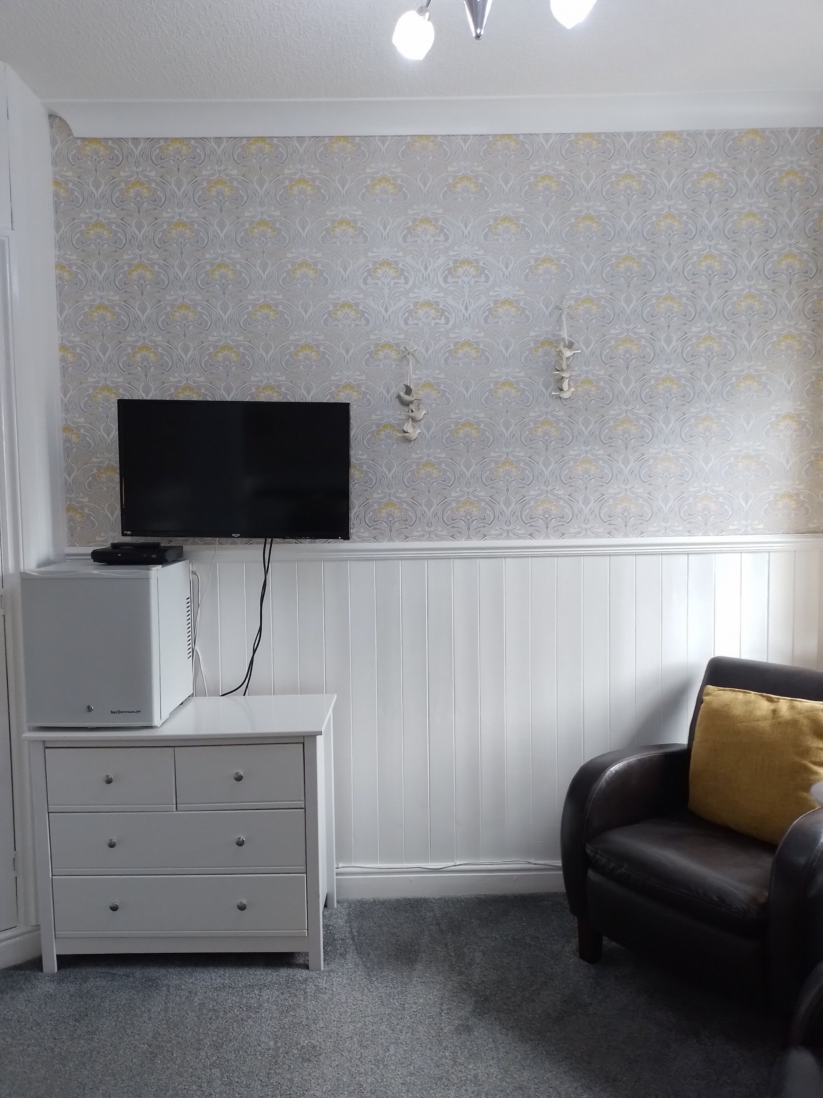 A room with a white dresser, a flat-screen TV mounted on the wall, a black leather chair with a yellow pillow, and a patterned wallpaper.
