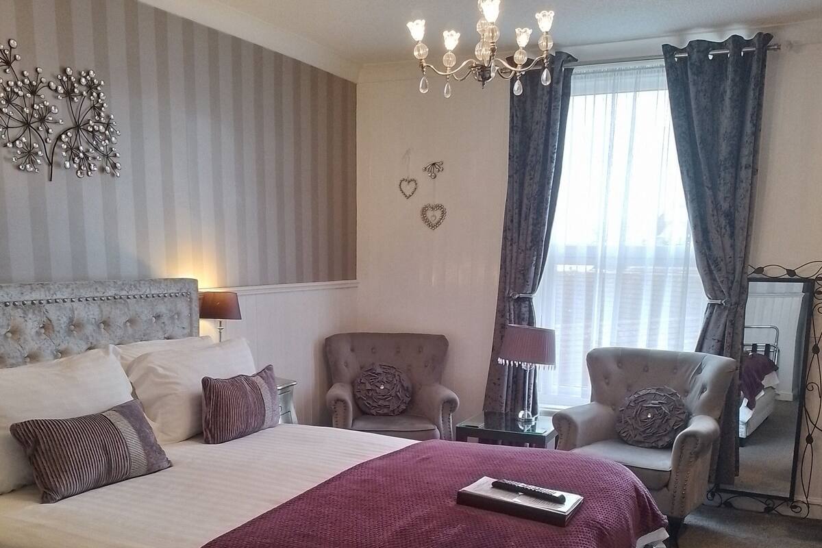 A well-appointed bedroom with a large bed, a chandelier, a sitting area with chairs, and a view of the outdoors through curtains.