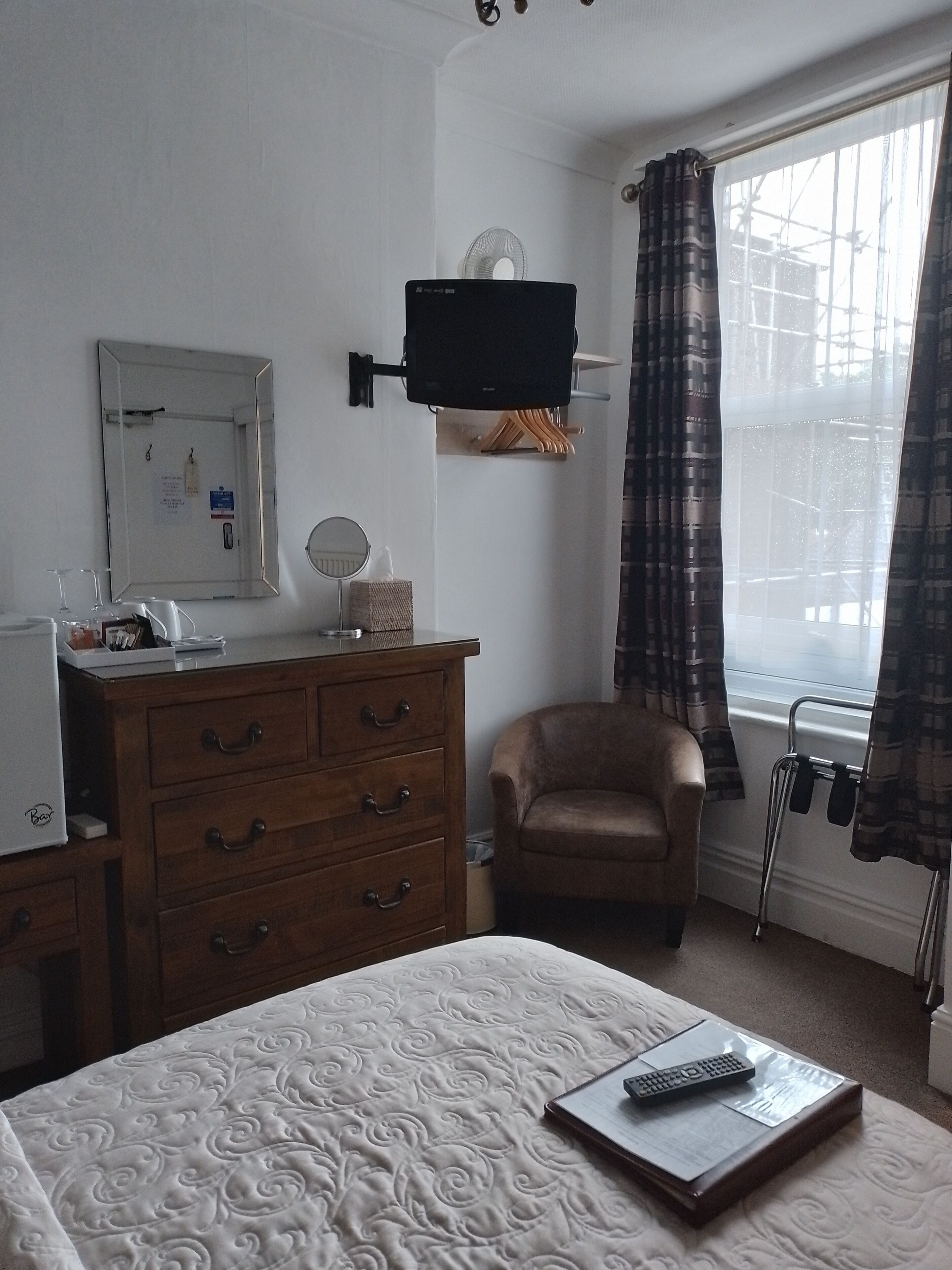 Ein Hotelzimmer mit Bett, einem hölzernen Schminktisch, einem Sessel, einem an der Wand montierten Fernseher und einem Fenster mit Vorhängen.