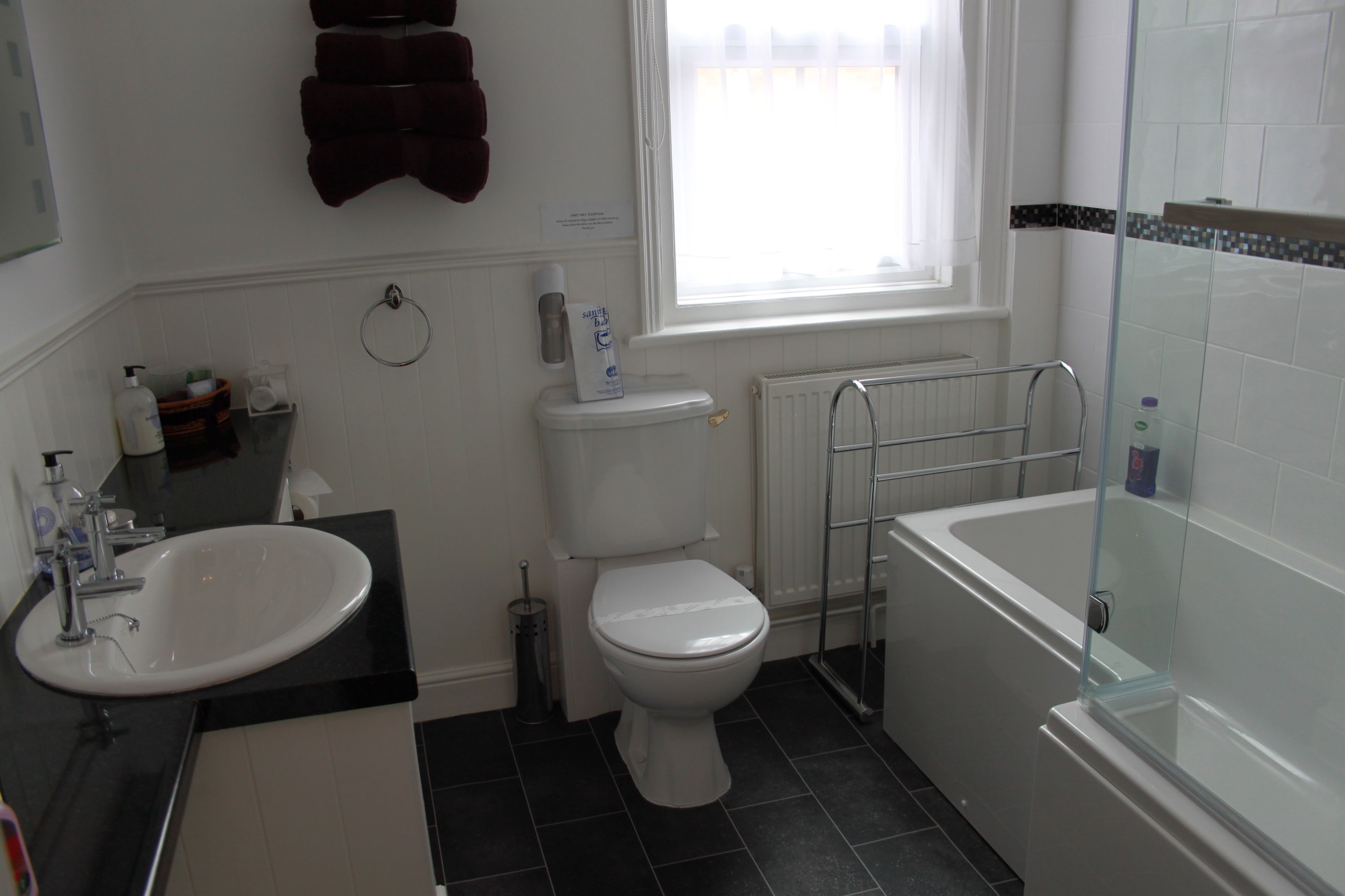 A bathroom with a white sink, toilet, and bathtub. There is a window with white curtains, a towel rack, and a soap dispenser on the wall.