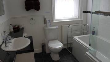A bathroom with a white sink, toilet, and bathtub. There is a window with white curtains, a towel rack, and a soap dispenser on the wall.