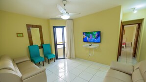 A living room with beige sofas, teal chairs, a television, and a ceiling fan. - Edifício Londres (Guarapari)