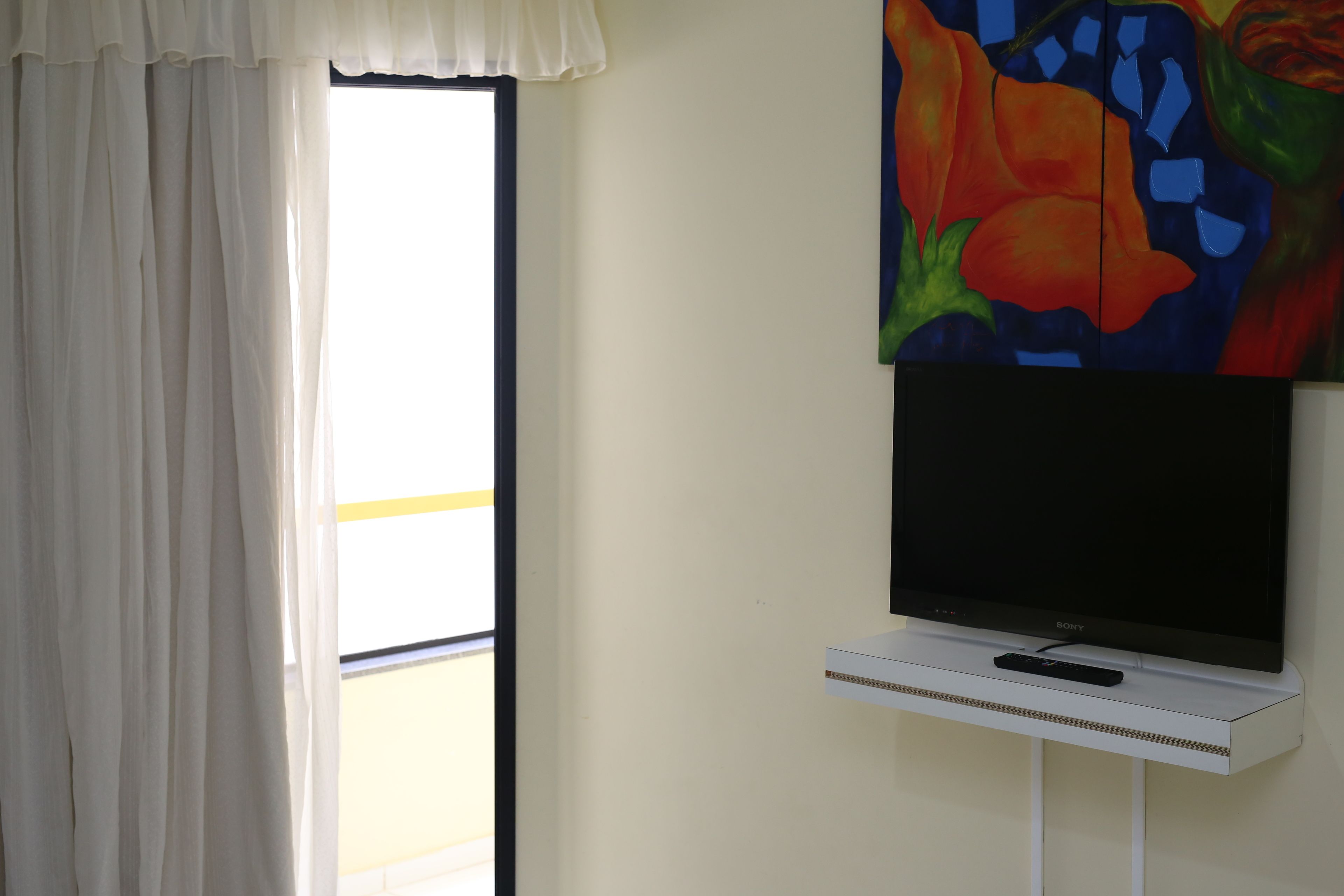 A room with a television on a white stand, a window with white curtains, and a colorful painting on the wall.