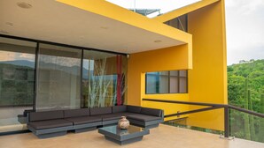 A modern outdoor seating area with a sofa and a coffee table, a glass wall with a view of greenery, and a yellow building facade.