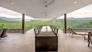 A long dining table with chairs, a ceiling fan, and a view of green hills.