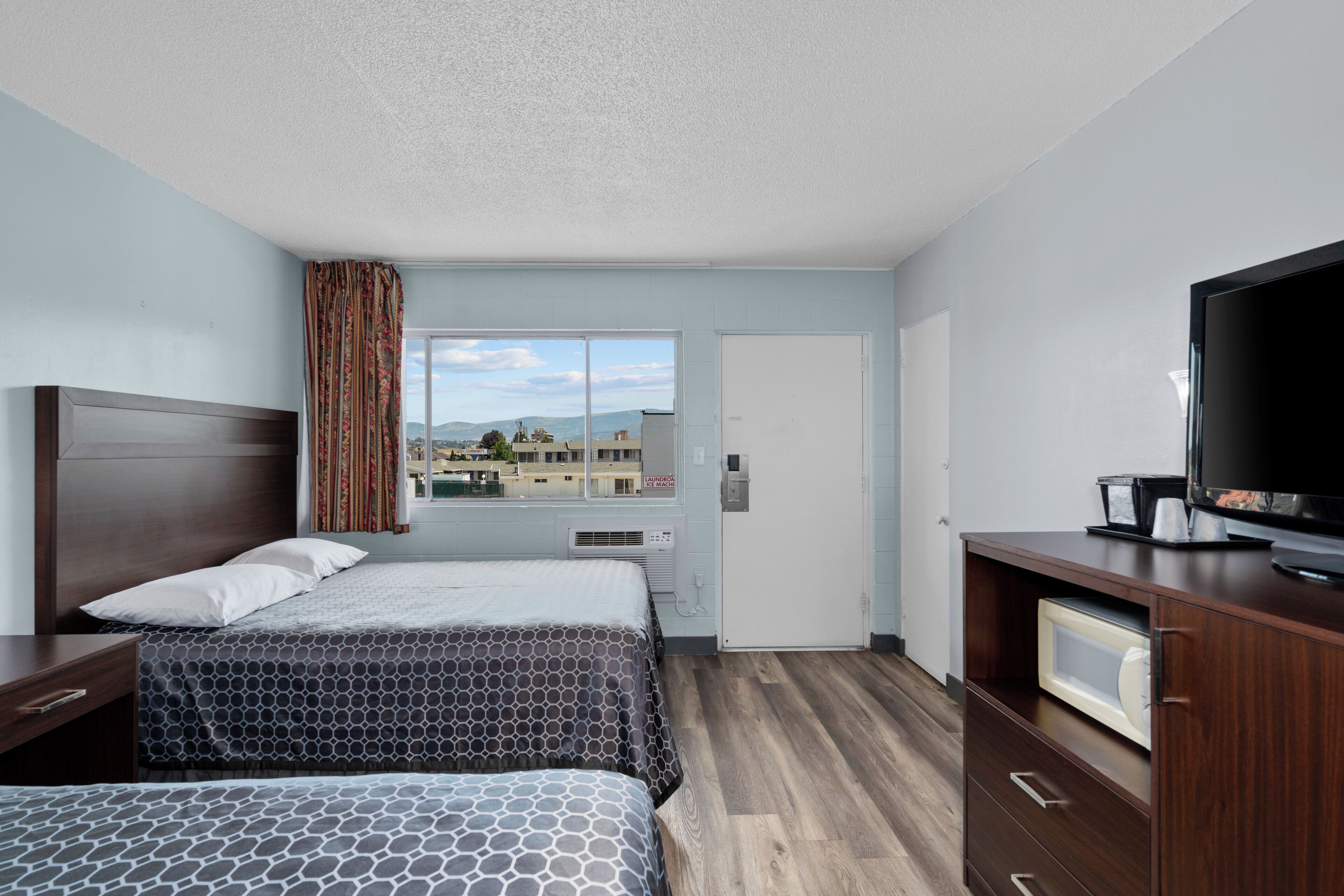 Une chambre d’hôtel avec deux lits, une télévision, une commode et une fenêtre donnant sur des immeubles et des montagnes.