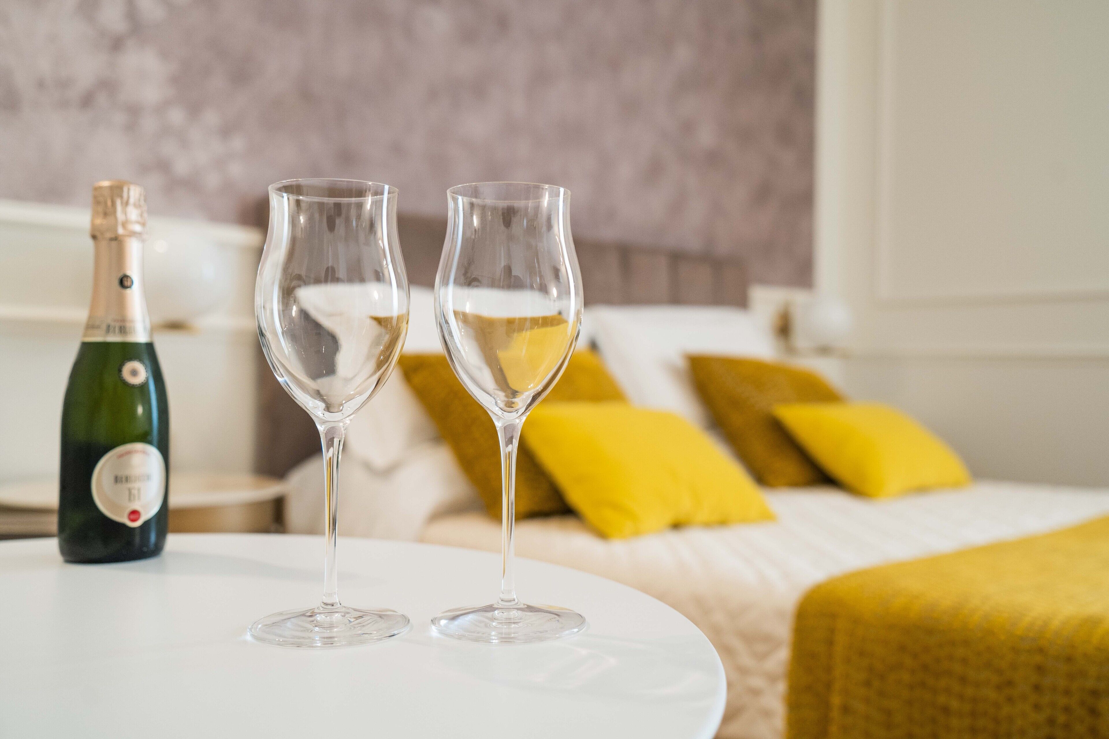 Una cama con almohadas amarillas y una manta blanca, una botella de champán y dos copas de vino vacías sobre una mesa blanca.