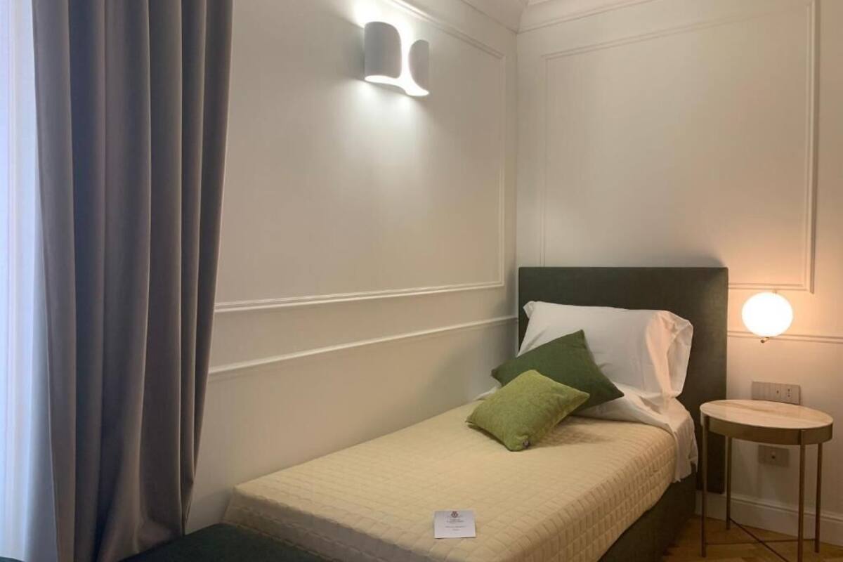 Ein kleines, ordentlich bezogenes Bett mit einem grünen und einem weißen Kopfkissen, ein Nachttisch mit einer Lampe sowie ein zur Seite gezogen Vorhang.