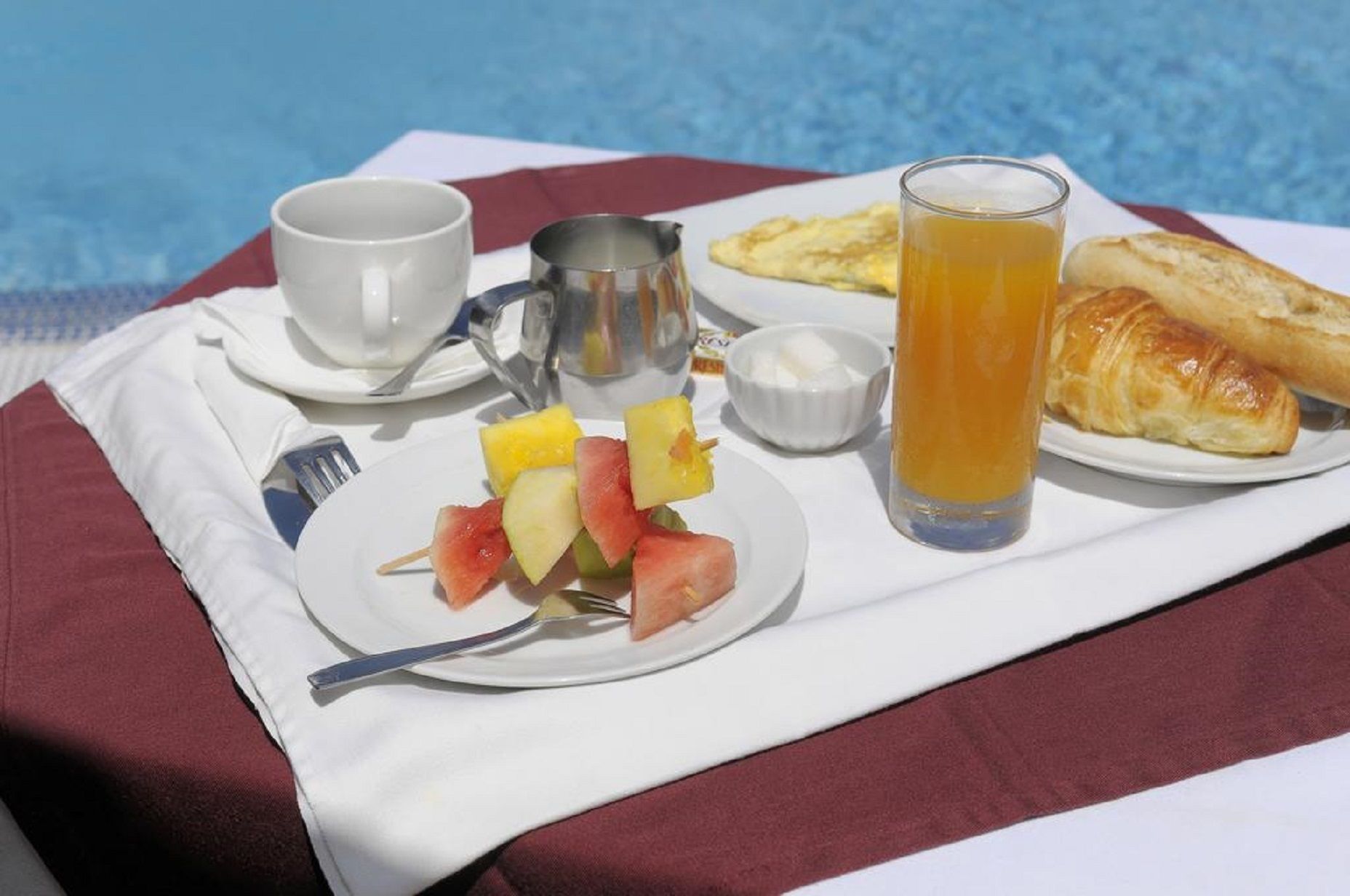 Bandeja de desayuno con brochetas de fruta, un croissant, un vaso de zumo de naranja, una taza y un recipiente pequeño con mantequilla sobre un mantel blanco junto a una piscina.