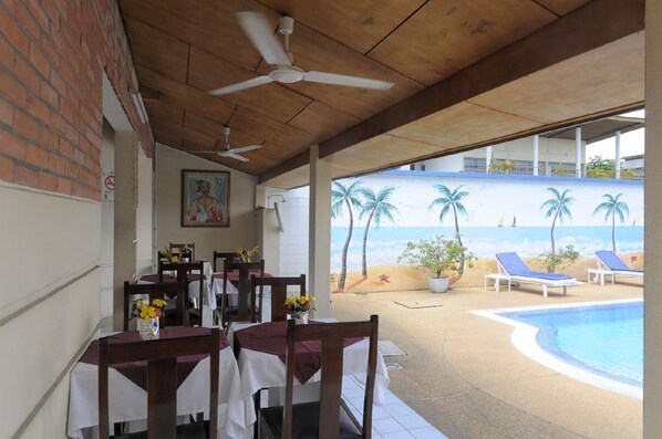 A covered outdoor dining area with tables and chairs, a ceiling fan, a mural of a beach scene, and a pool with lounge chairs.