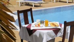 Área de refeições junto à piscina, com uma mesa posta para o pequeno-almoço, incluindo croissants, fruta e sumo.