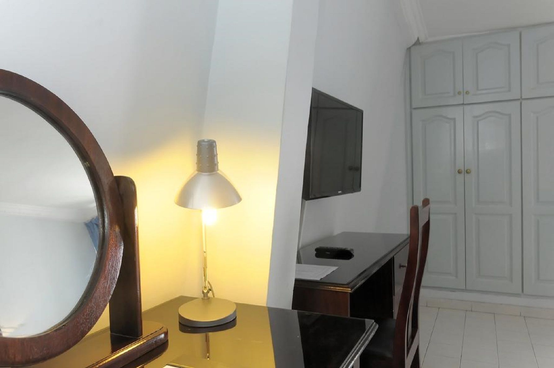 A room with a desk, a round mirror, a lamp, a TV mounted on the wall, and a wardrobe.