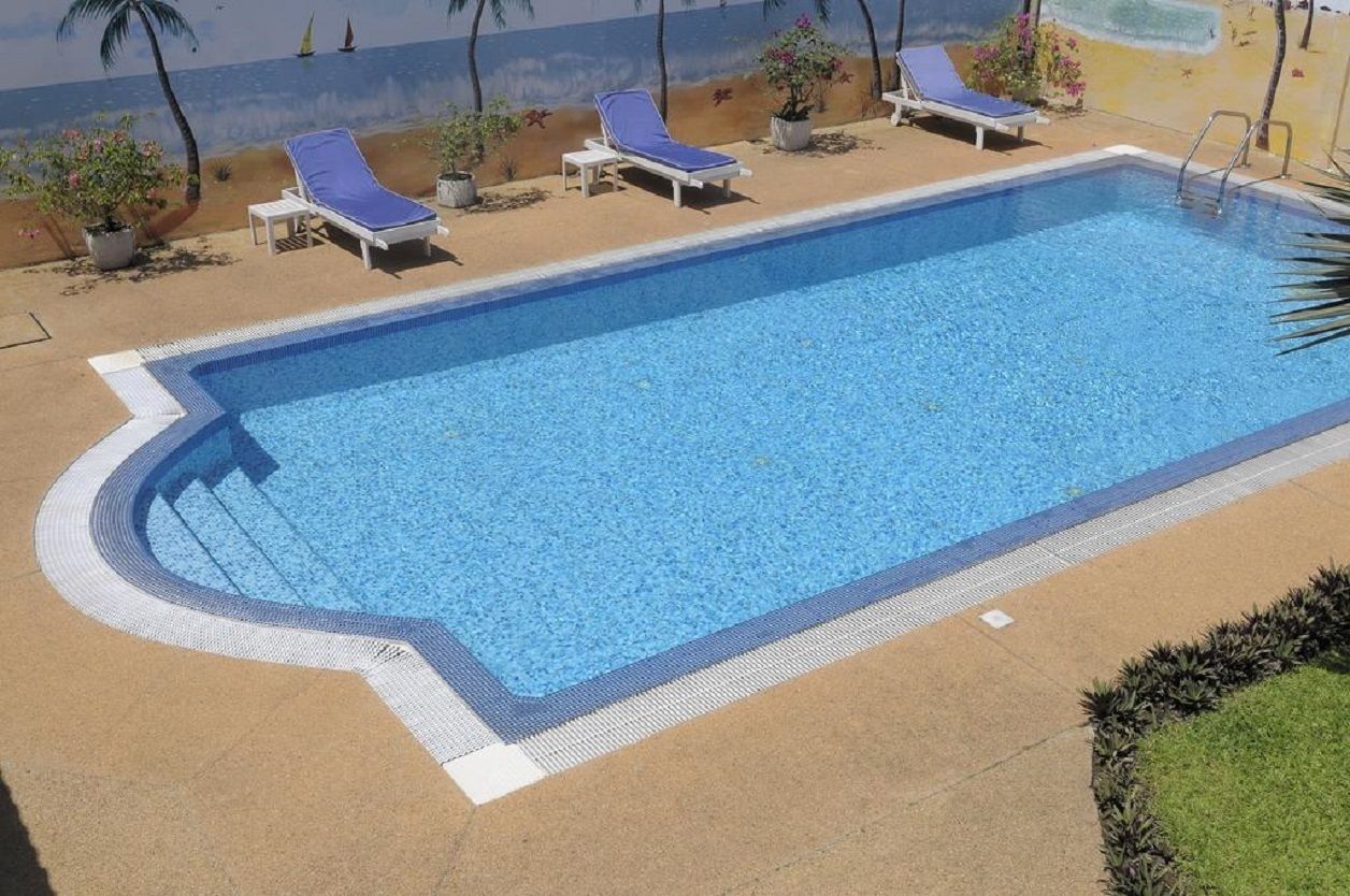 A rectangular swimming pool with a blue interior, surrounded by a sandy deck. There are three lounge chairs with blue cushions and white frames.