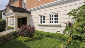 Uma casa de dois andares com fachada de tijolos, varanda coberta e um jardim bem cuidado com arbustos e flores variadas.