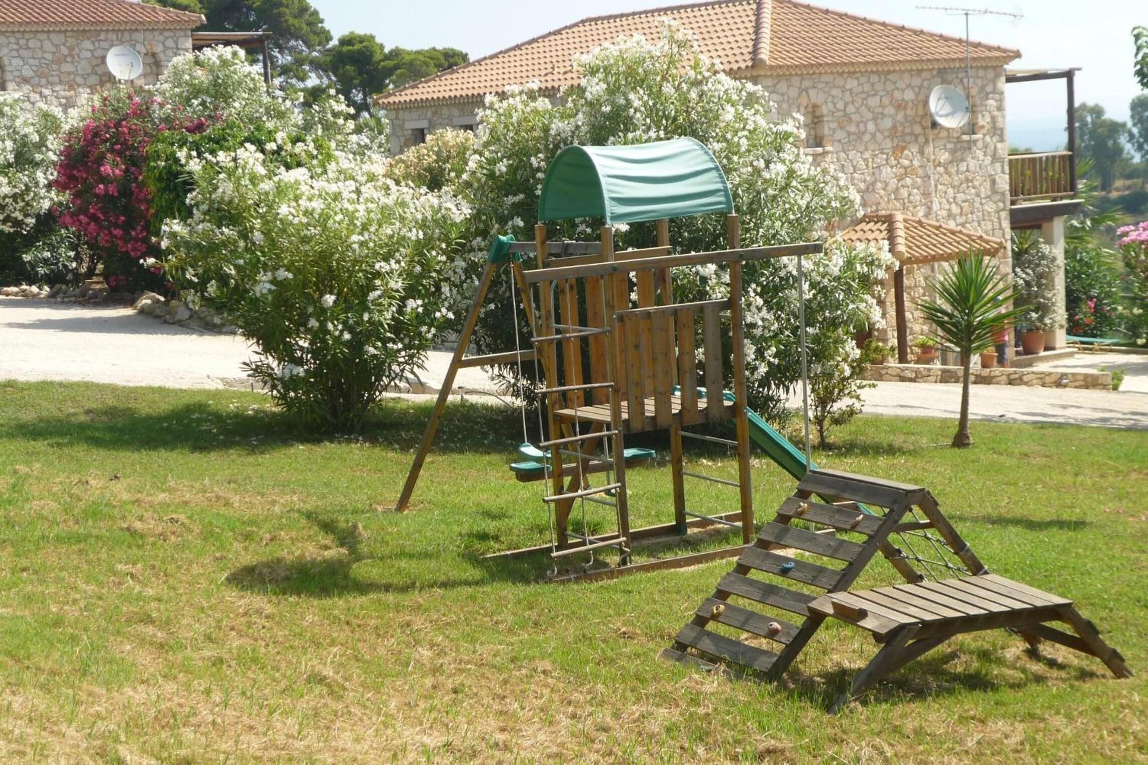 A playground with a swing set and a slide, surrounded by lush greenery and a stone building in the background.