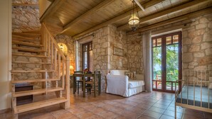 A rustic interior with a stone wall, wooden staircase, a dining area with a table and chairs, a white sofa, and a view through a glass door. - Myrties stone houses (Zakynthos)