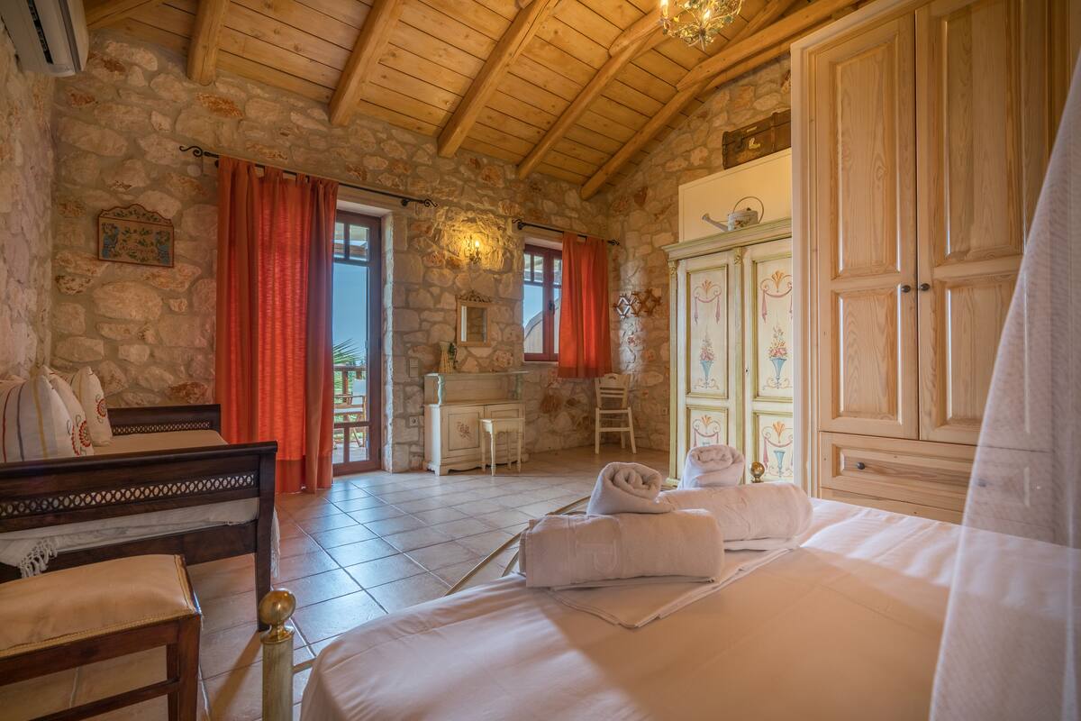 A bedroom with a stone wall, wooden ceiling, a bed with white linens, a dresser, and a wardrobe.