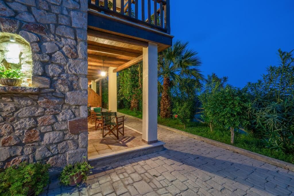 A stone building with a balcony, a dining area with outdoor furniture, and a well-maintained garden with palm trees.