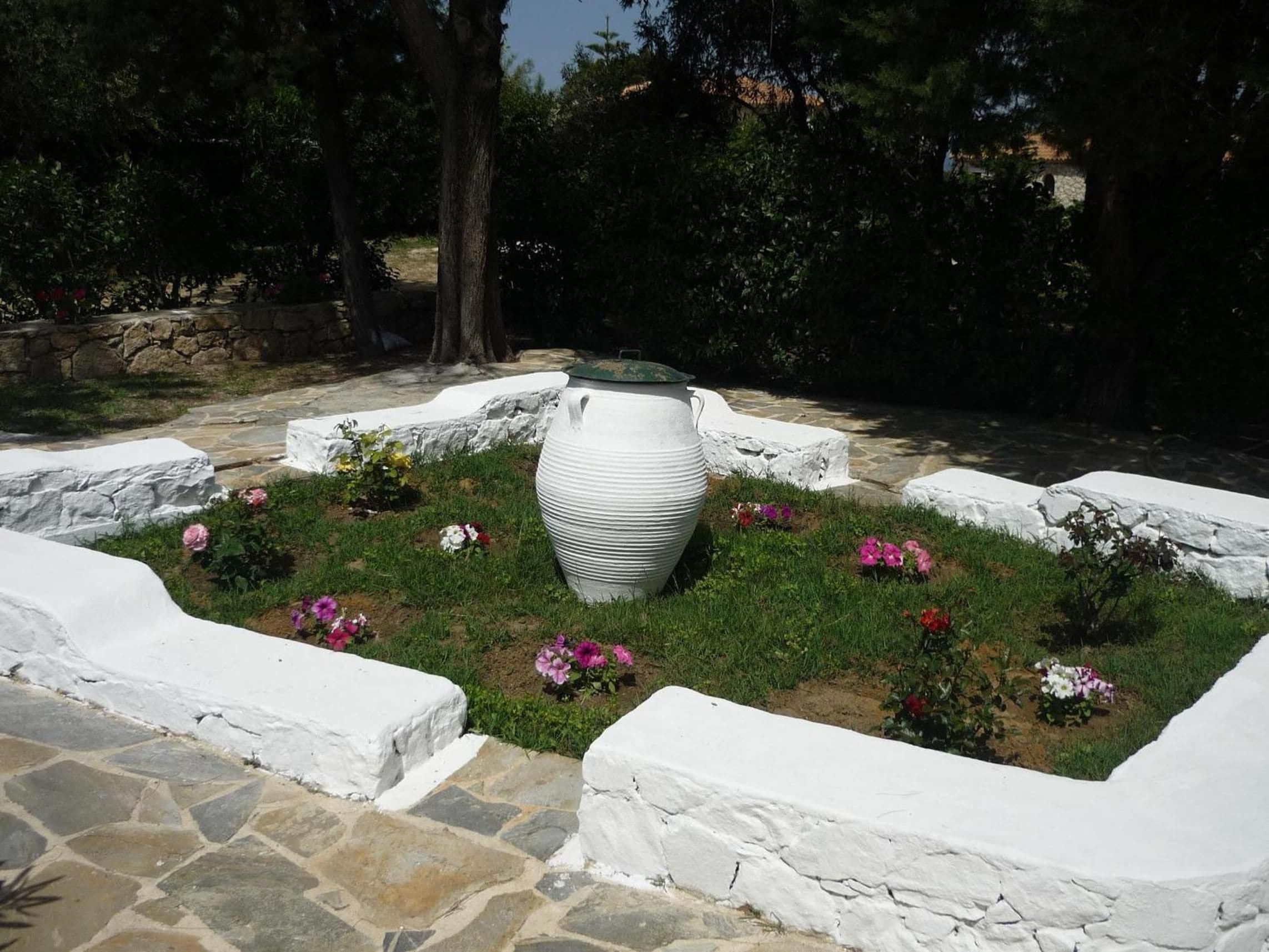 Un jardín con una urna grande de color blanco, rodeado de muros de piedra blanca y diversas flores.