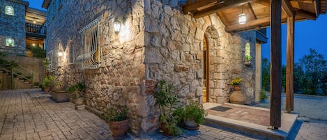 A stone building with a wooden pergola entrance, potted plants, and a cobblestone pathway.