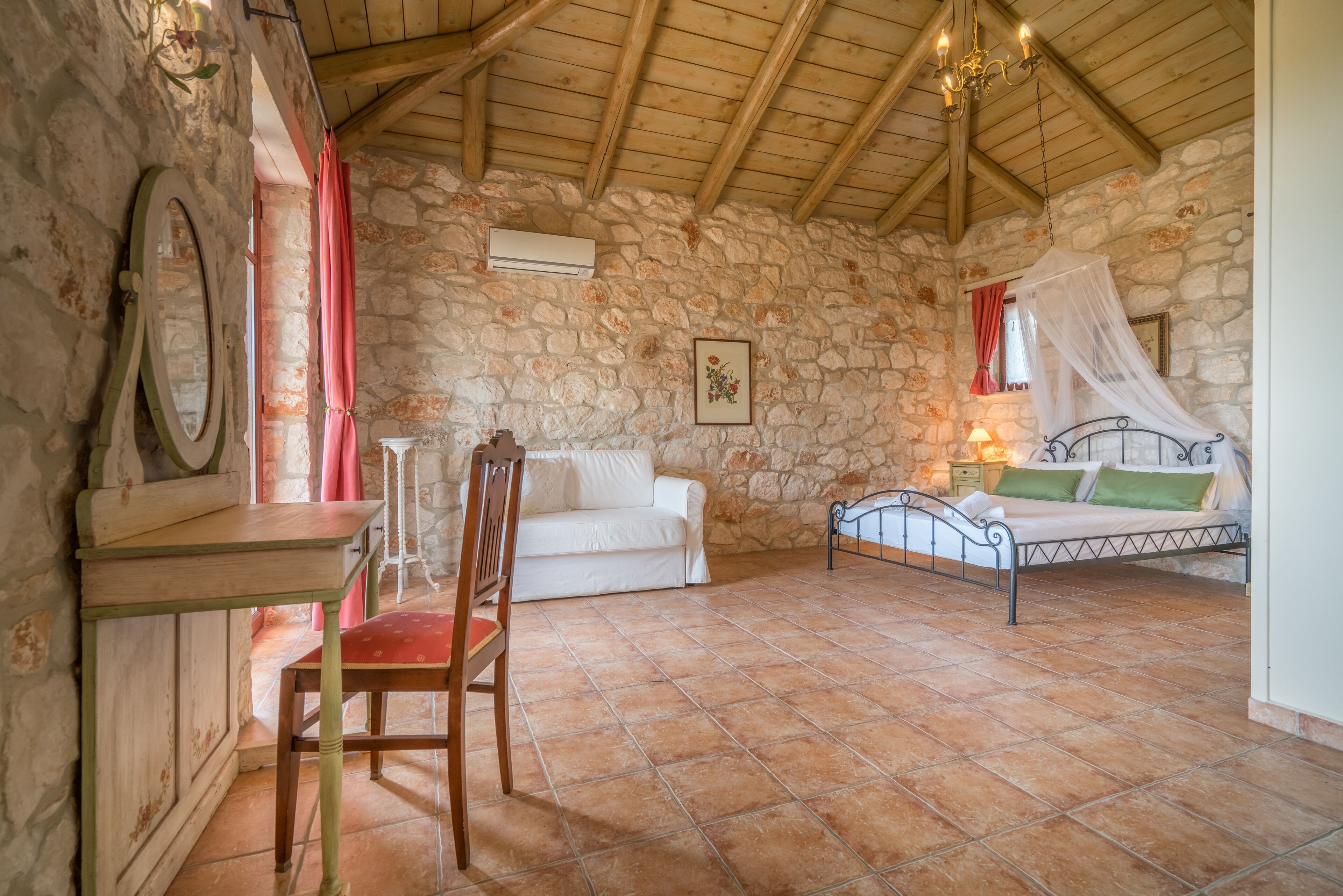 A rustic room with a stone wall, a wooden ceiling, a bed with a canopy, a white sofa, a chair, and a small table.