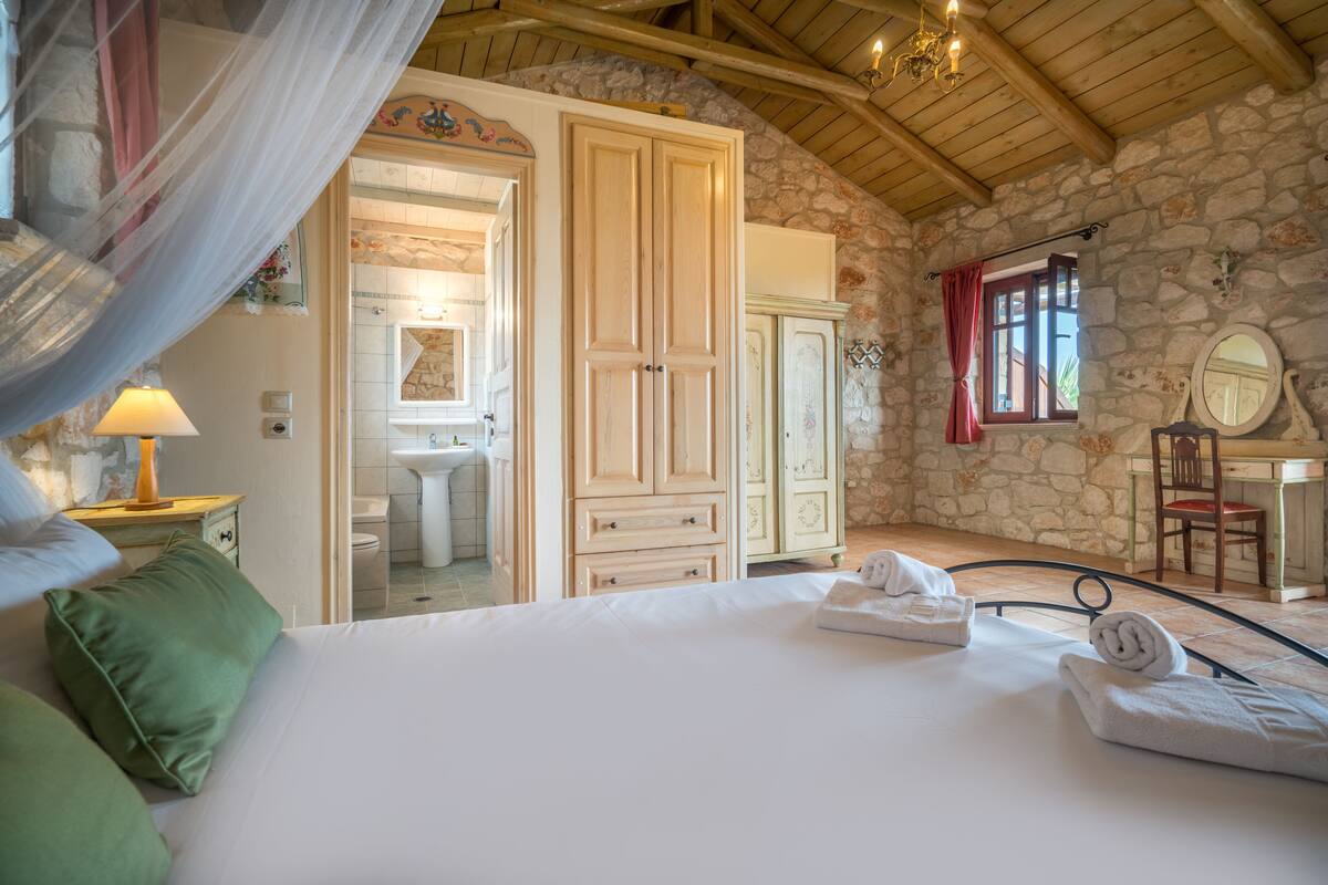 A bedroom with a large bed, a wooden wardrobe, a stone wall, and a bathroom visible through an open door.