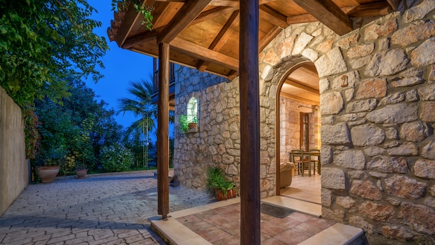 A stone-walled outdoor patio with a wooden pergola, potted plants, and a view of the surrounding greenery. - Myrties stone houses (Zakynthos)