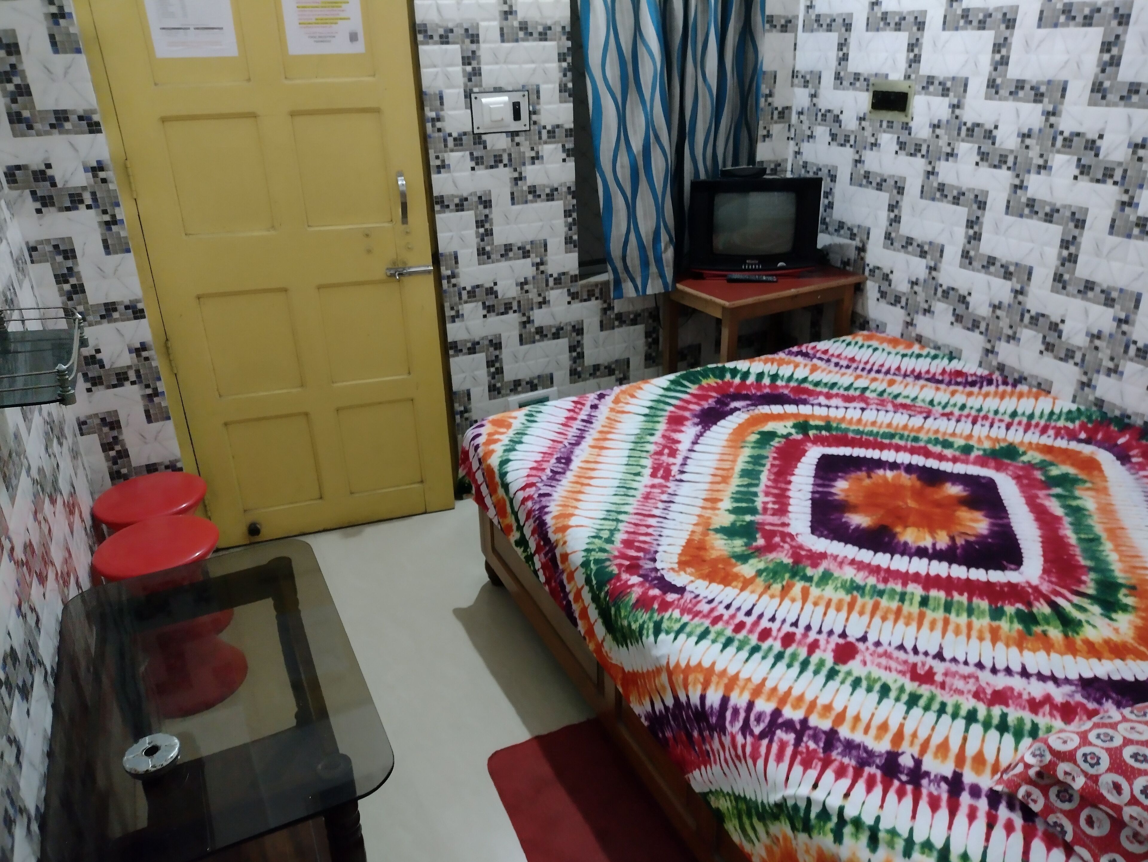 Een kamer met een gele deur, een bed met een kleurrijke deken, een kleine televisie op een houten tafel en een rode kruk.