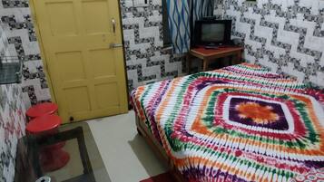 Een kamer met een gele deur, een bed met een kleurrijke deken, een kleine televisie op een houten tafel en een rode kruk.