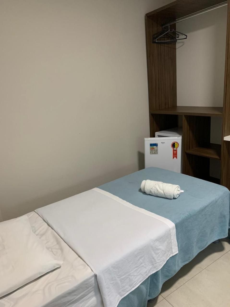 Een eenpersoonsbed met een wit laken en een blauwe deken, een kleine witte koelkast en een houten garderobe met spiegel.